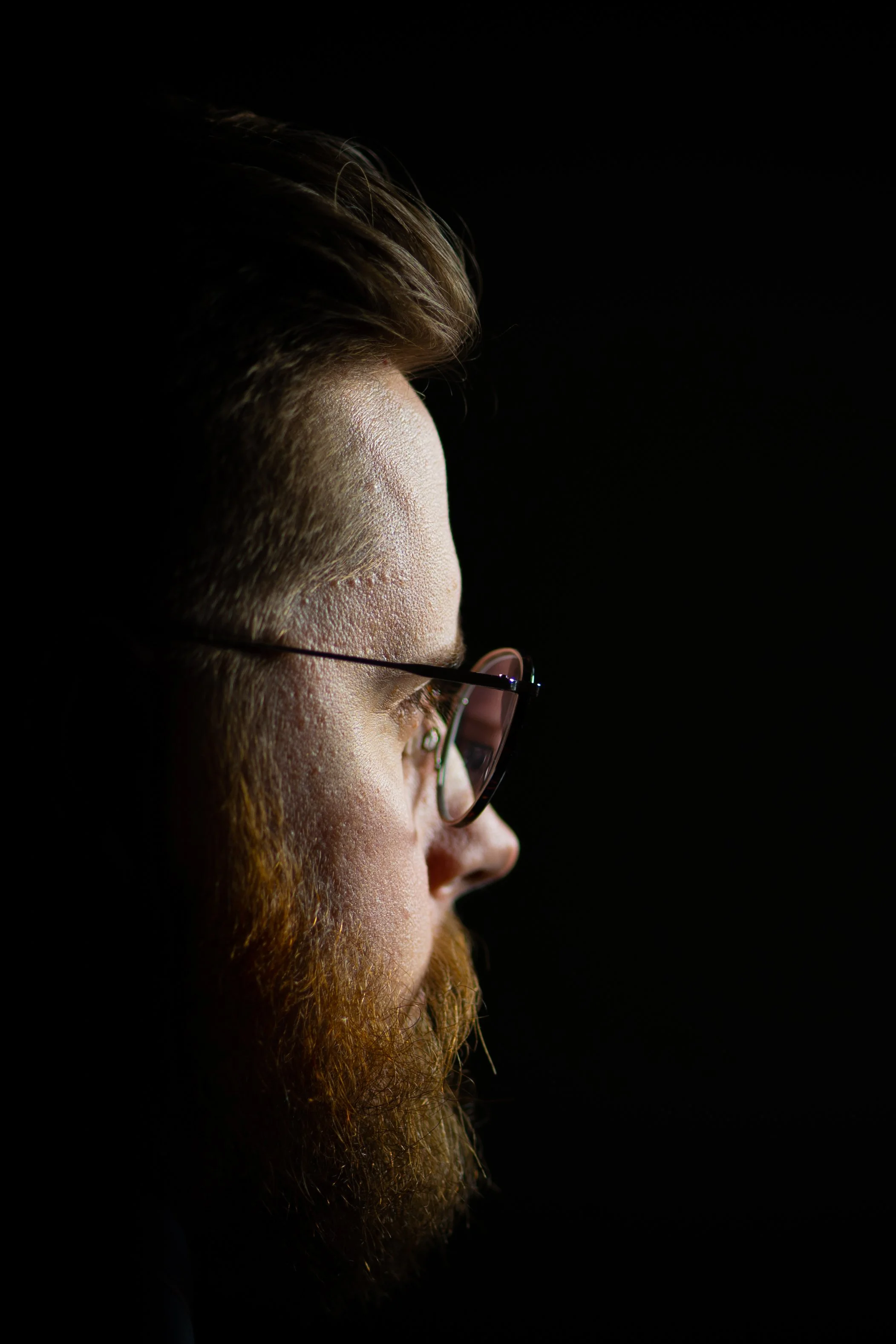 Close-up profile of a man with glasses and a red beard, looking to the right against a black background.