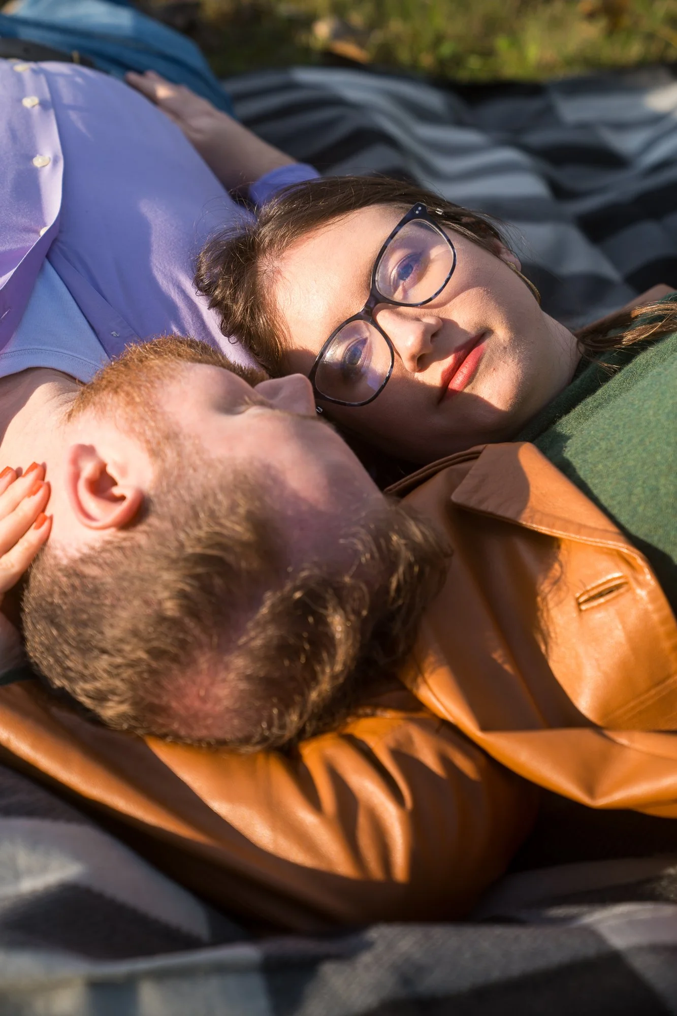 A couple lying on the grass outdoors, gazing at each other, with the woman wearing glasses and the man with a beard.