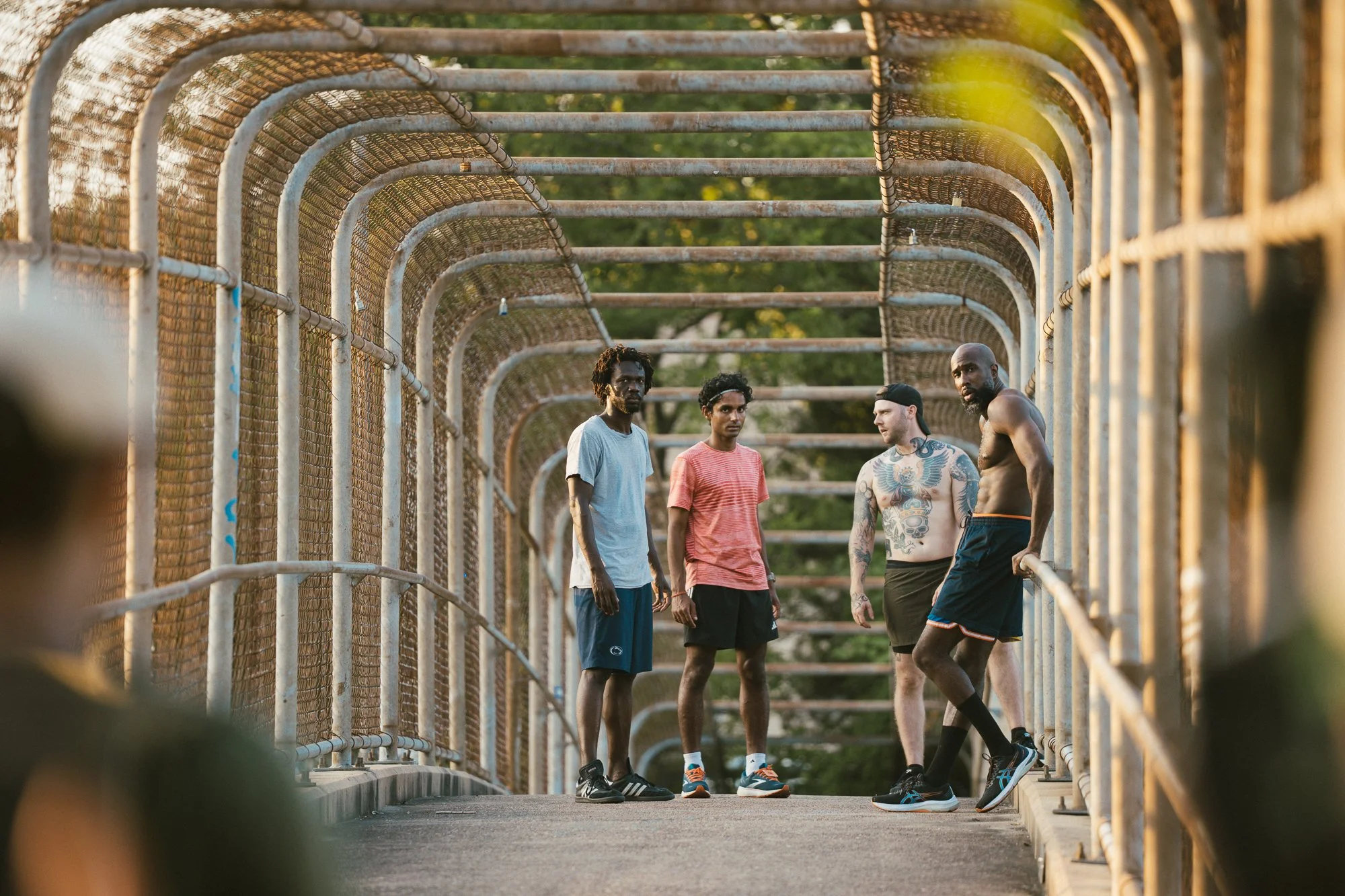 Four men standing and talking on a footbridge with metal railings and a mesh covering, bathed in warm sunlight.
