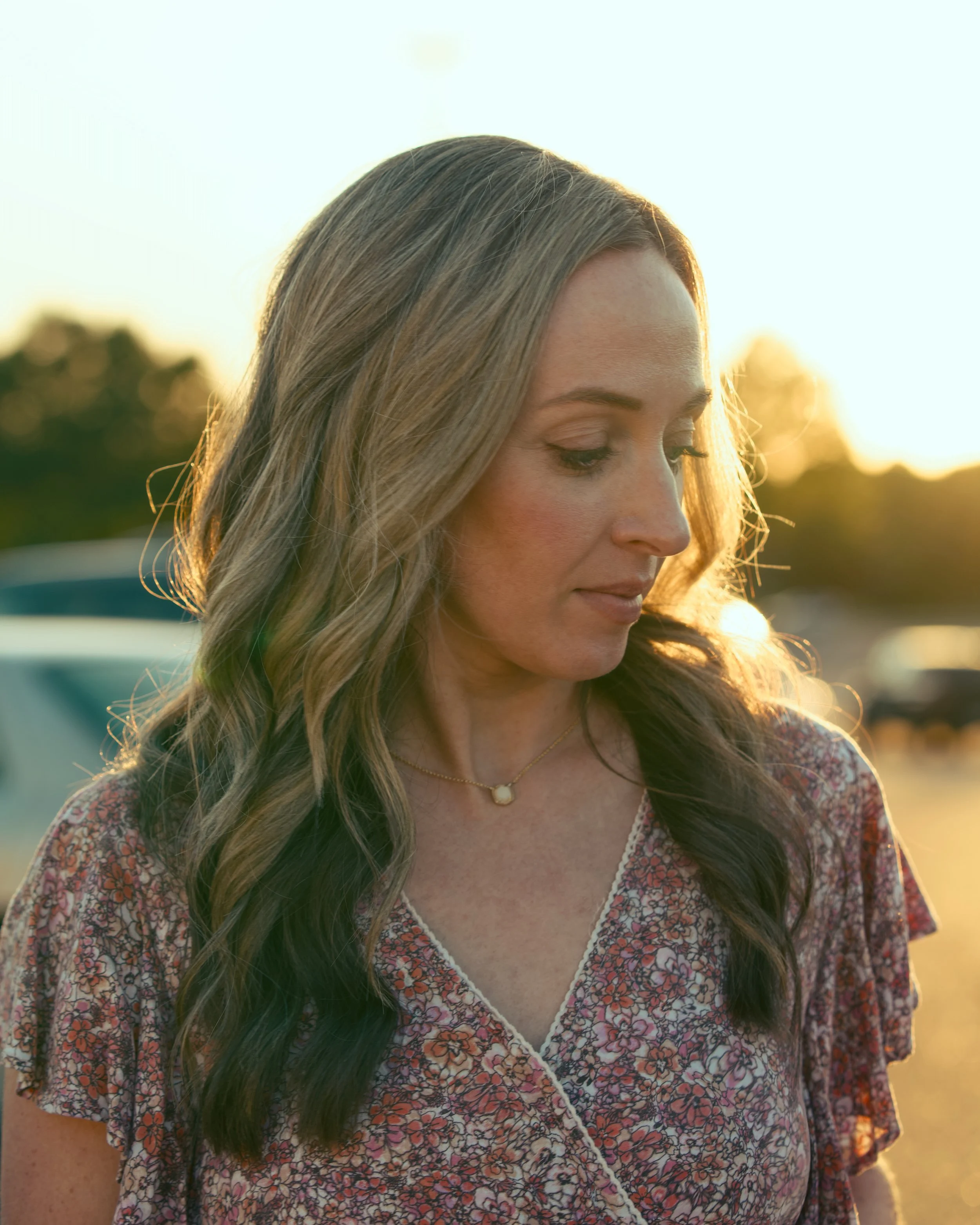 Close-up of a woman with long, wavy hair, wearing a floral dress and a pearl necklace, standing outdoors during sunset.
