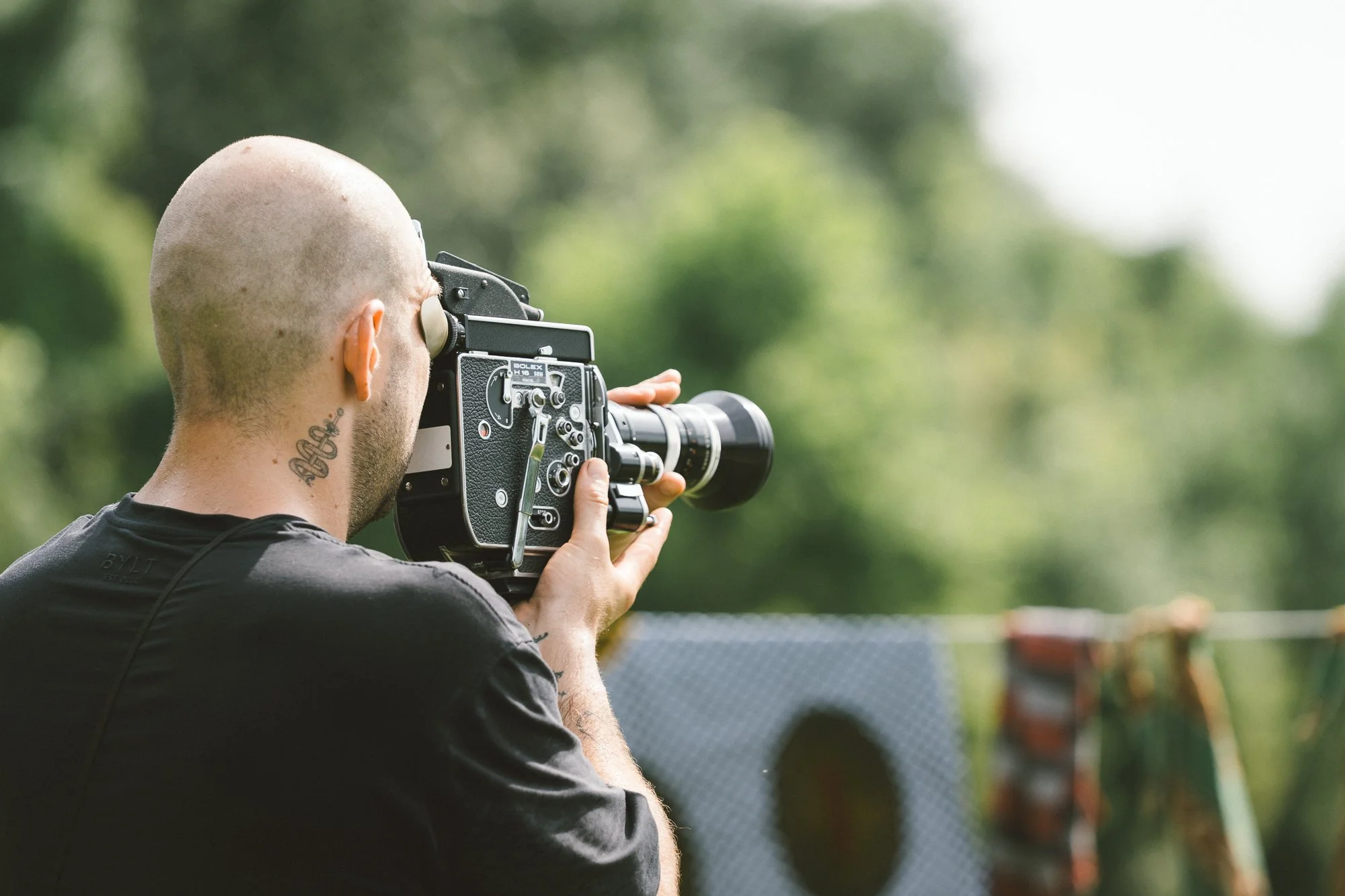 A man with a shaved head and tattoo on neck using a film camera outdoors