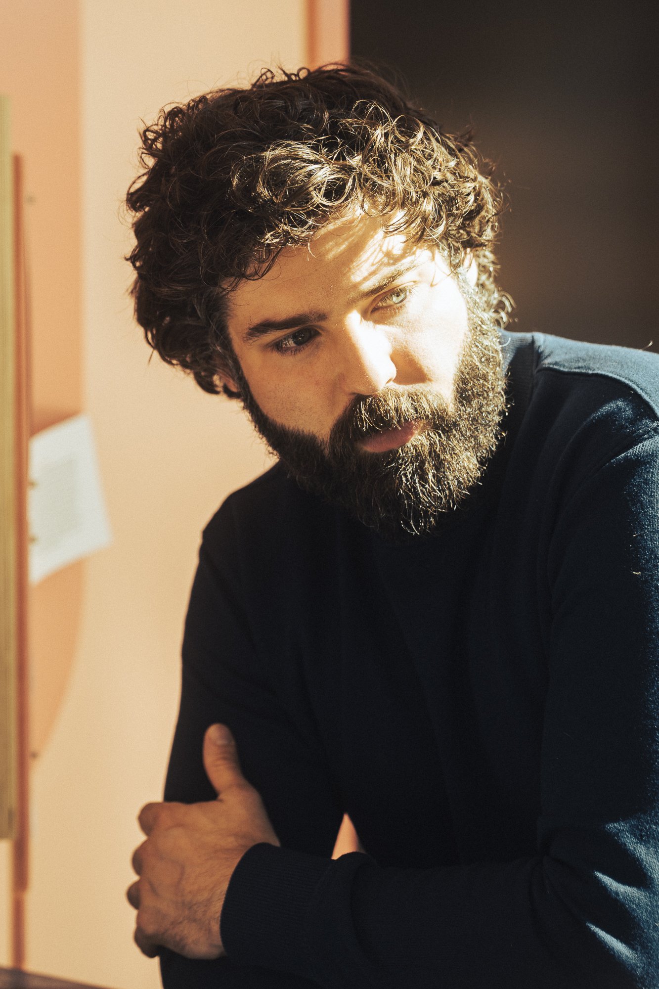 A man with curly hair and a beard looking down, wearing a black shirt, with his arms crossed, indoors with sunlight on his face.