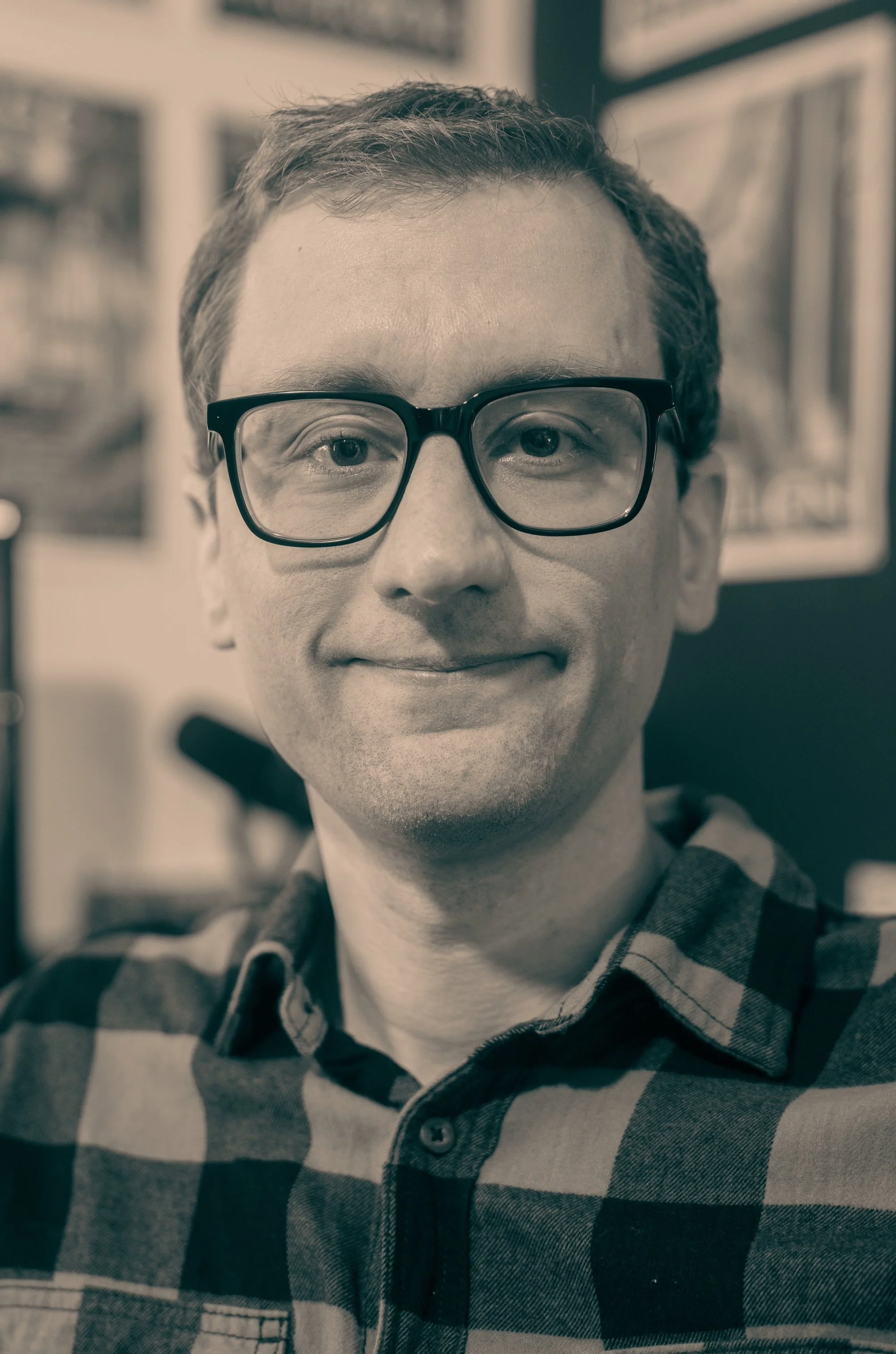 A man with glasses smiling, wearing a checkered shirt, indoors with framed pictures on the wall behind him.