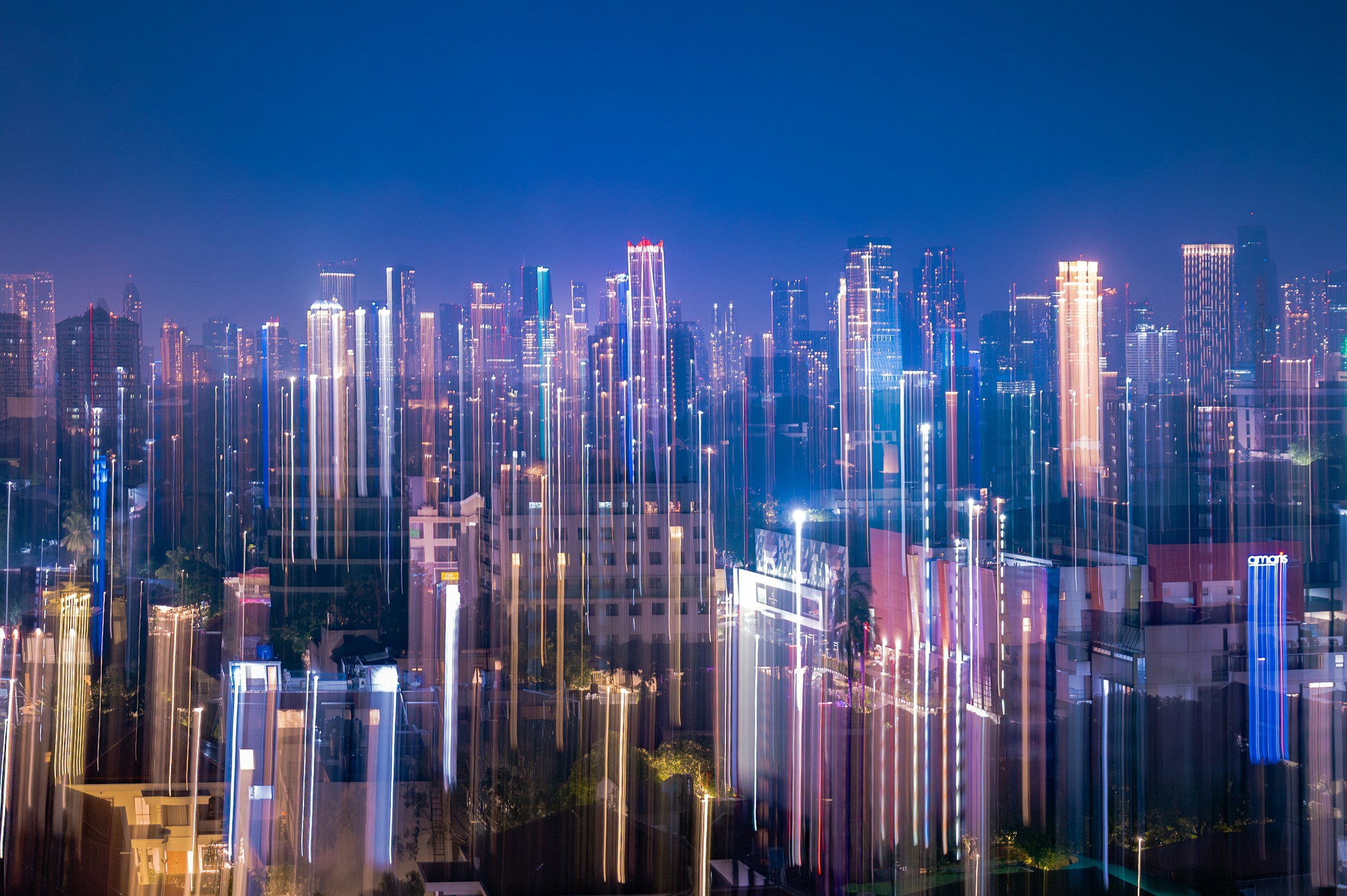 Nighttime city skyline with tall, illuminated skyscrapers and buildings, featuring light trails creating vertical streaks of light.