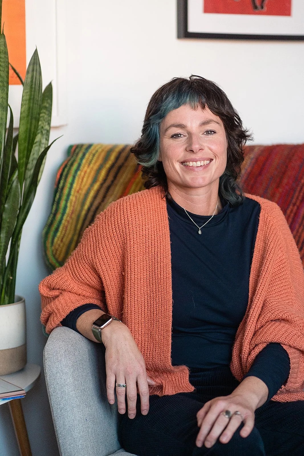 A woman with short dark hair and teal highlights smiling, sitting on a light gray armchair, with a multicolored blanket draped over a sofa behind her, a potted plant to her left, and artwork on the wall.