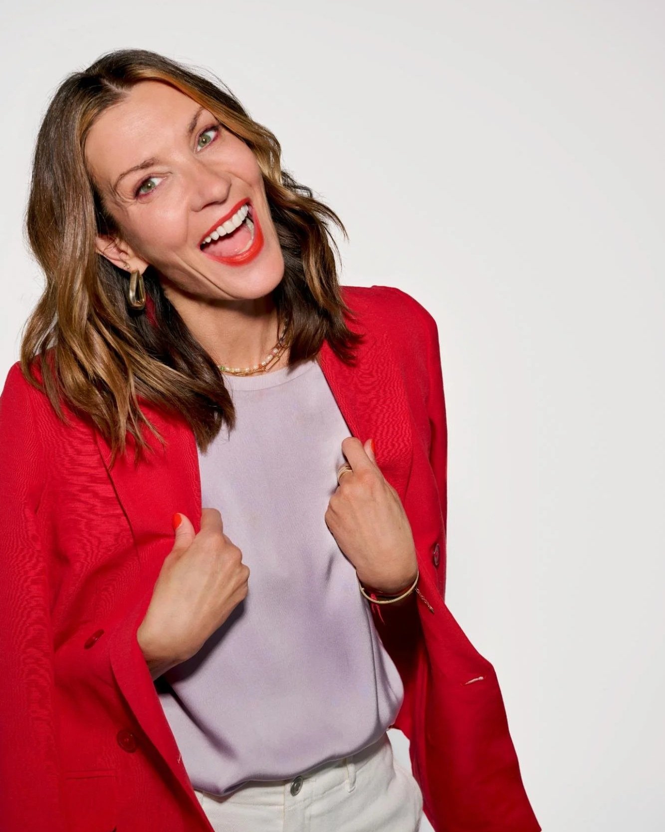 A woman with shoulder-length brown hair, wearing a red blazer over a light gray top, smiling broadly, holding her blazer lapels, against a plain white background.