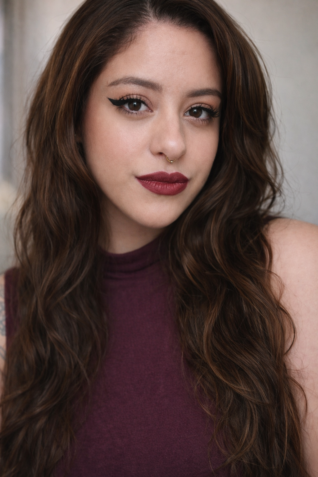A young woman with long wavy brown hair, pierced septum, wearing dark red lipstick, eye makeup, and a sleeveless dark purple top.