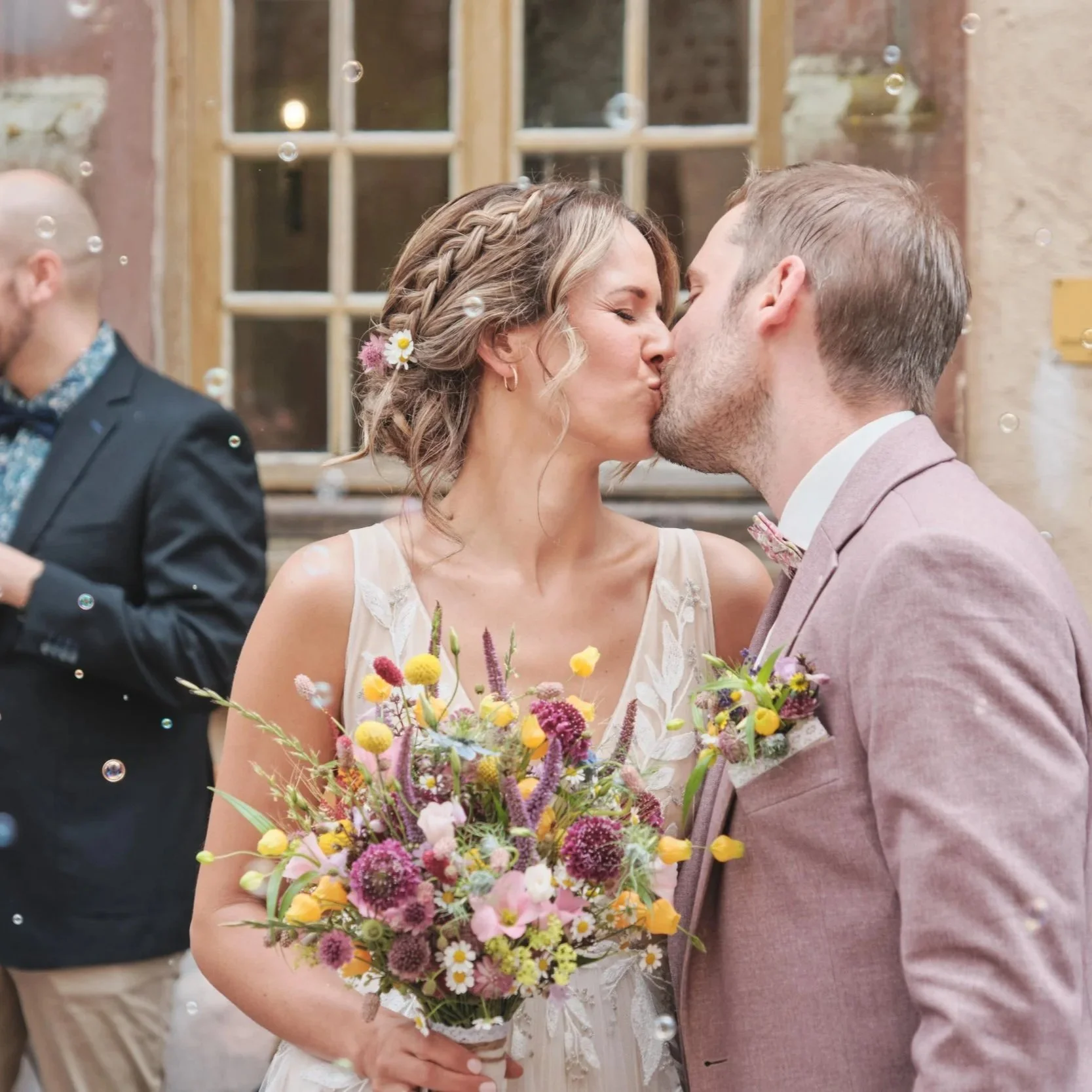 Ein Brautpaar küsst sich bei einer Hochzeit. Die Braut trägt ein weißes Kleid und hält einen bunten Blumenstrauß. Der Bräutigam trägt einen hellen Anzug mit Fliege. Im Hintergrund sind weitere Gäste und ein altes Gebäude sichtbar.