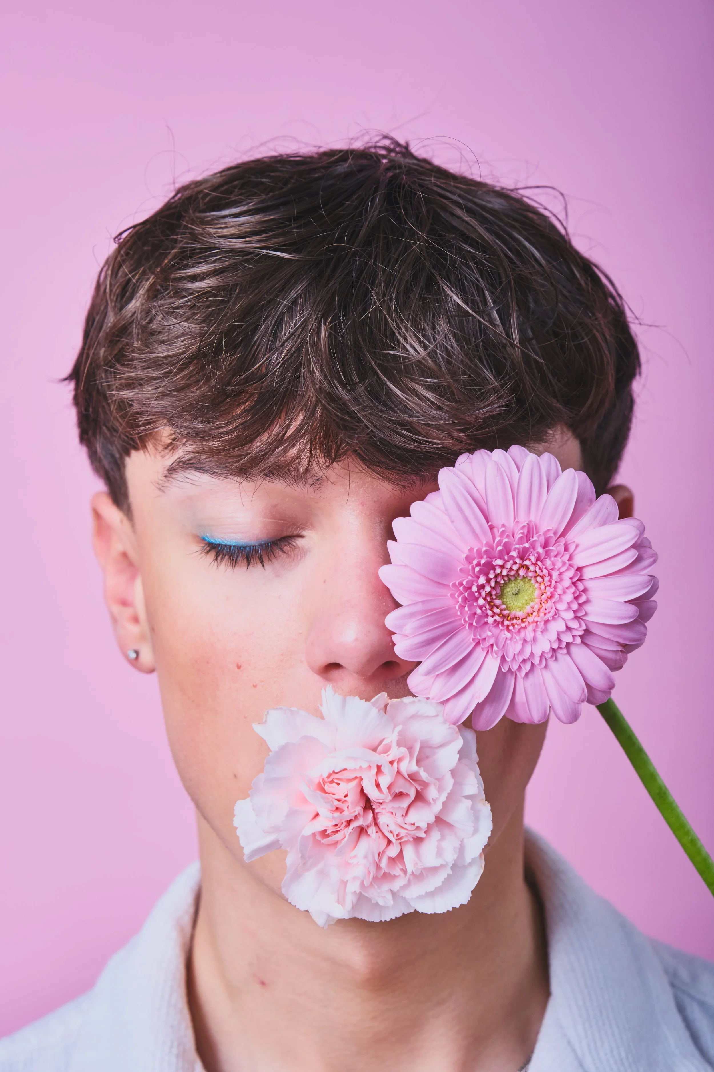 Porträt eines jungen Menschen mit geschlossenen Augen, der eine pinke und eine weiße Blume vor dem Gesicht hält, vor einem rosa Hintergrund.