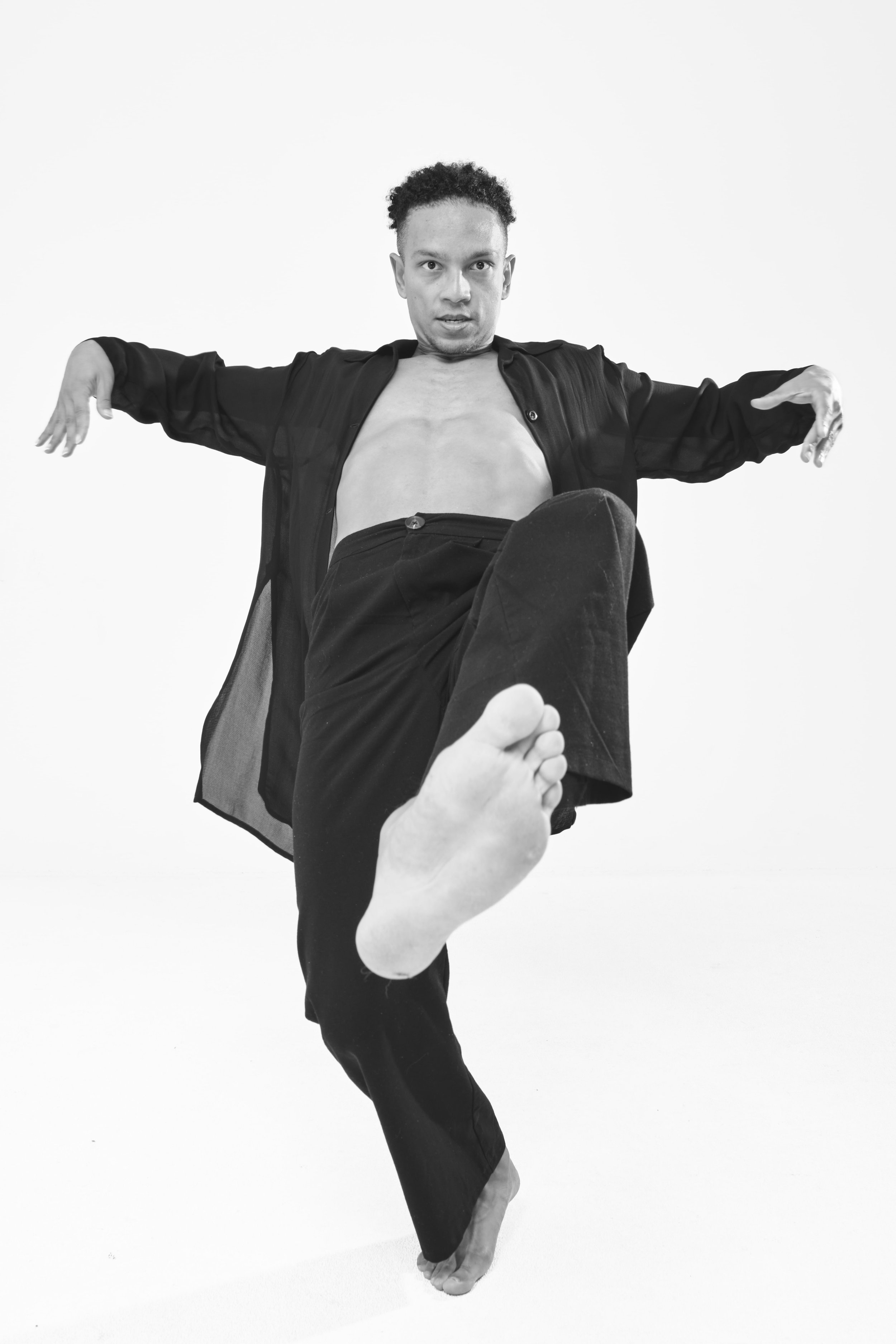 Schwarzweiß-Foto eines Mannes in einer Tanzpose, hebt ein Bein an, Arme ausgestreckt, trägt dunkle Kleidung und geöffnetes Hemd.