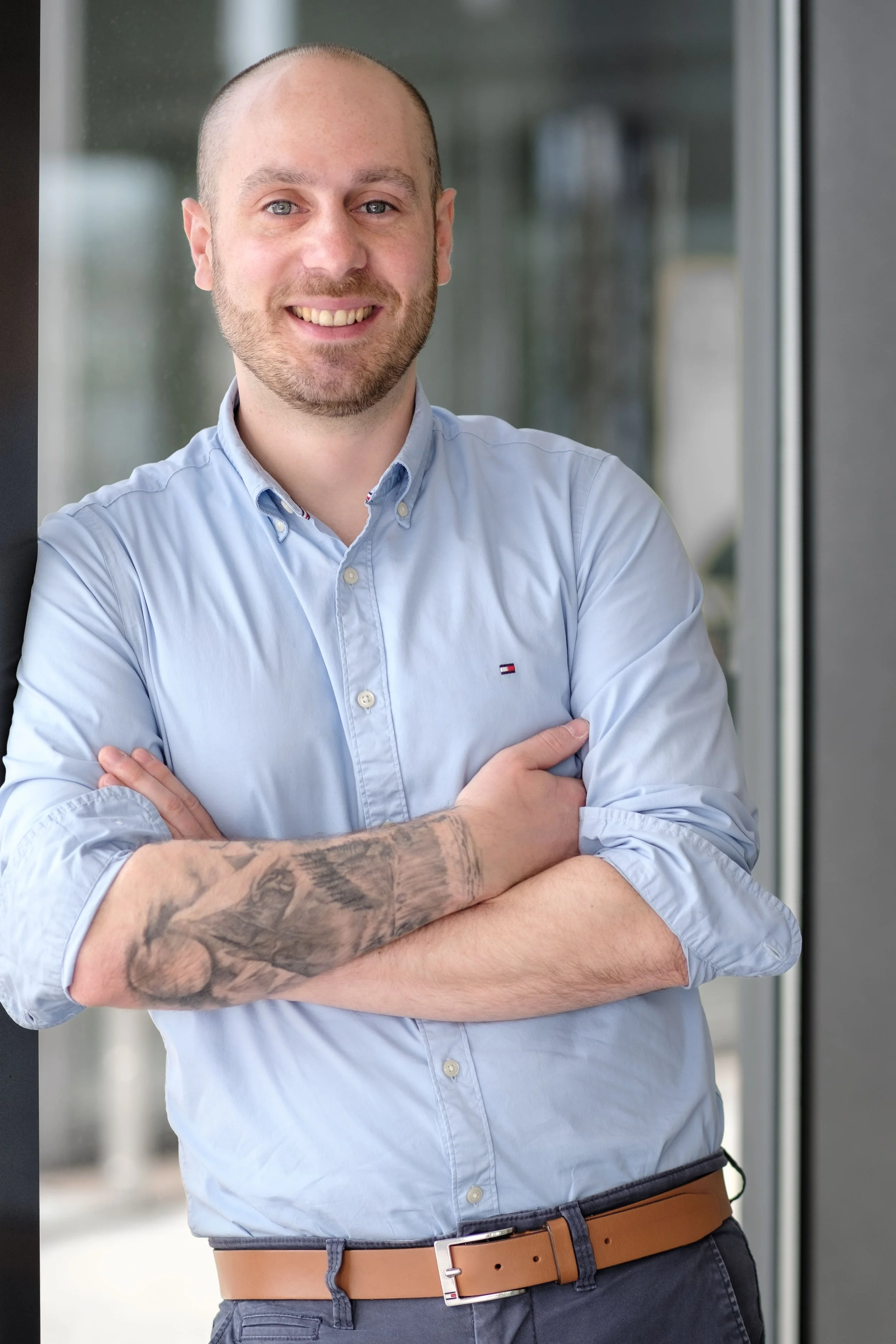 Ein lachender Mann mit einem kurz geschnittenen Bart und Tattoos an einem Arm steht mit verschränkten Armen vor einem Fenster in einem modernen Gebäude.