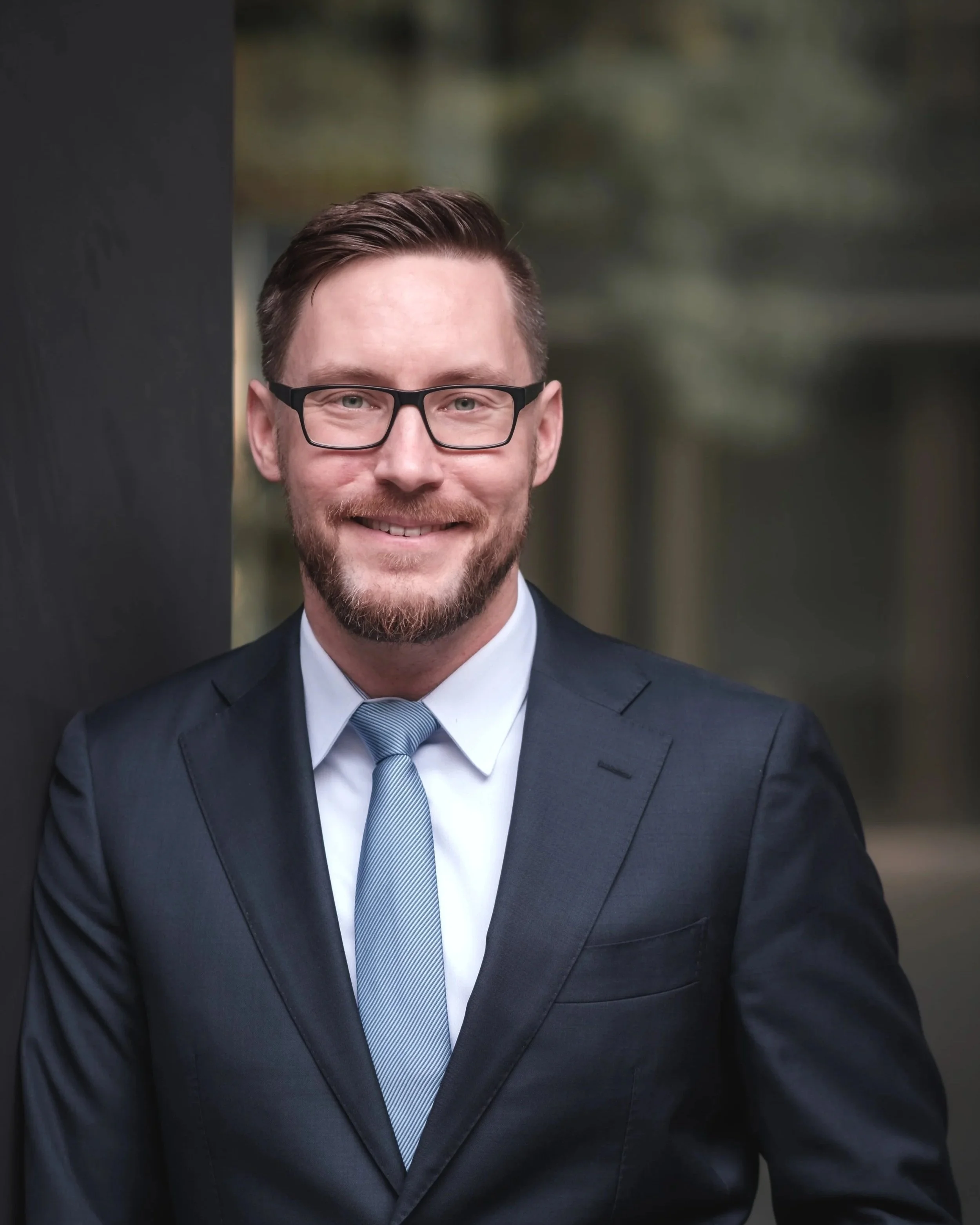 Businessportrait eines Mannes im dunklen Anzug, weißem Hemd und blauer Krawatte, trägt Brille und lächelt, steht vor einer dunklen Wand im Freien in Mainz.