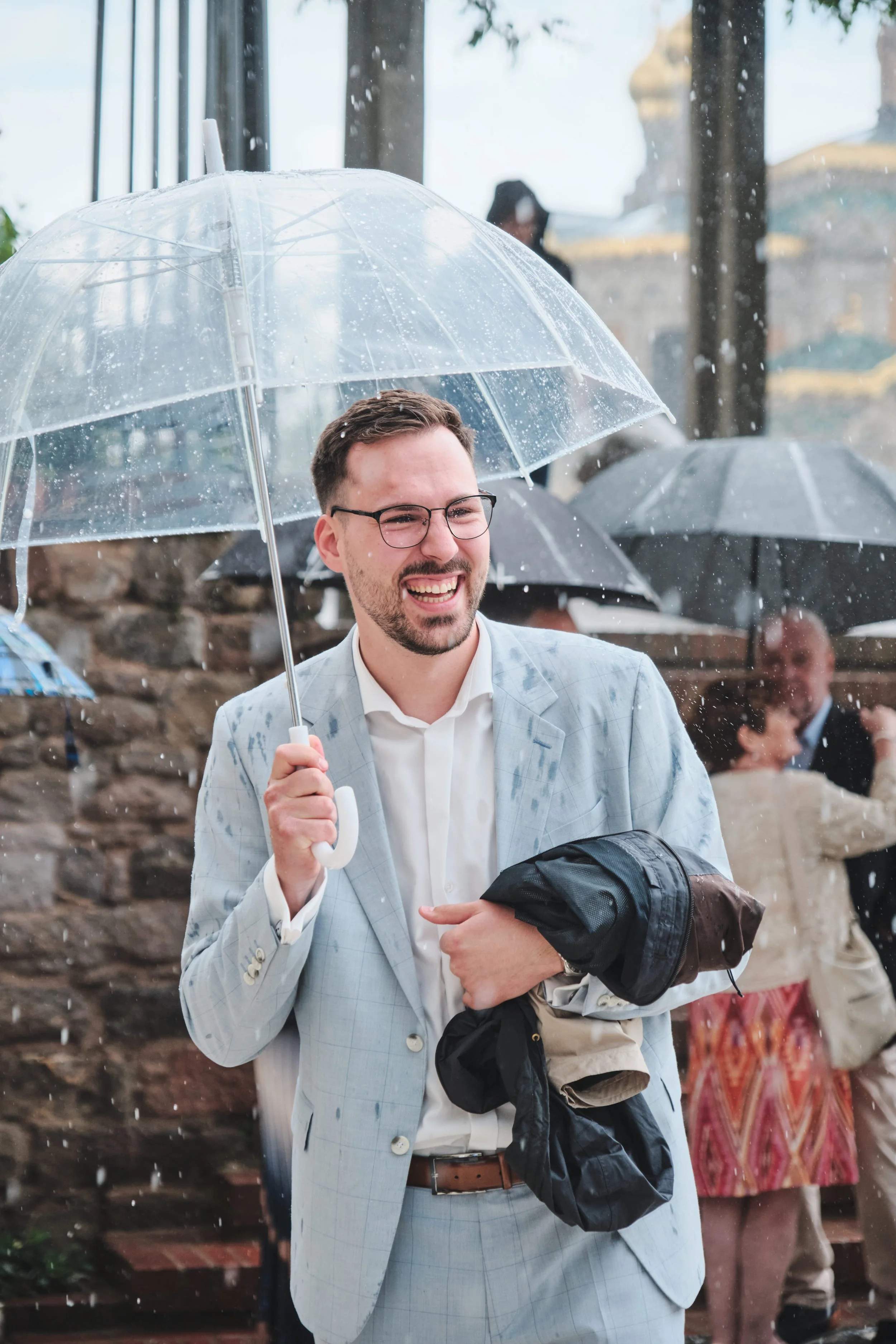 Der Bräutigam trägt einen grauen Anzug, hält einen transparenten Regenschirm und lacht im Regen, während andere Personen im Hintergrund ebenfalls Regenschirme halten.
