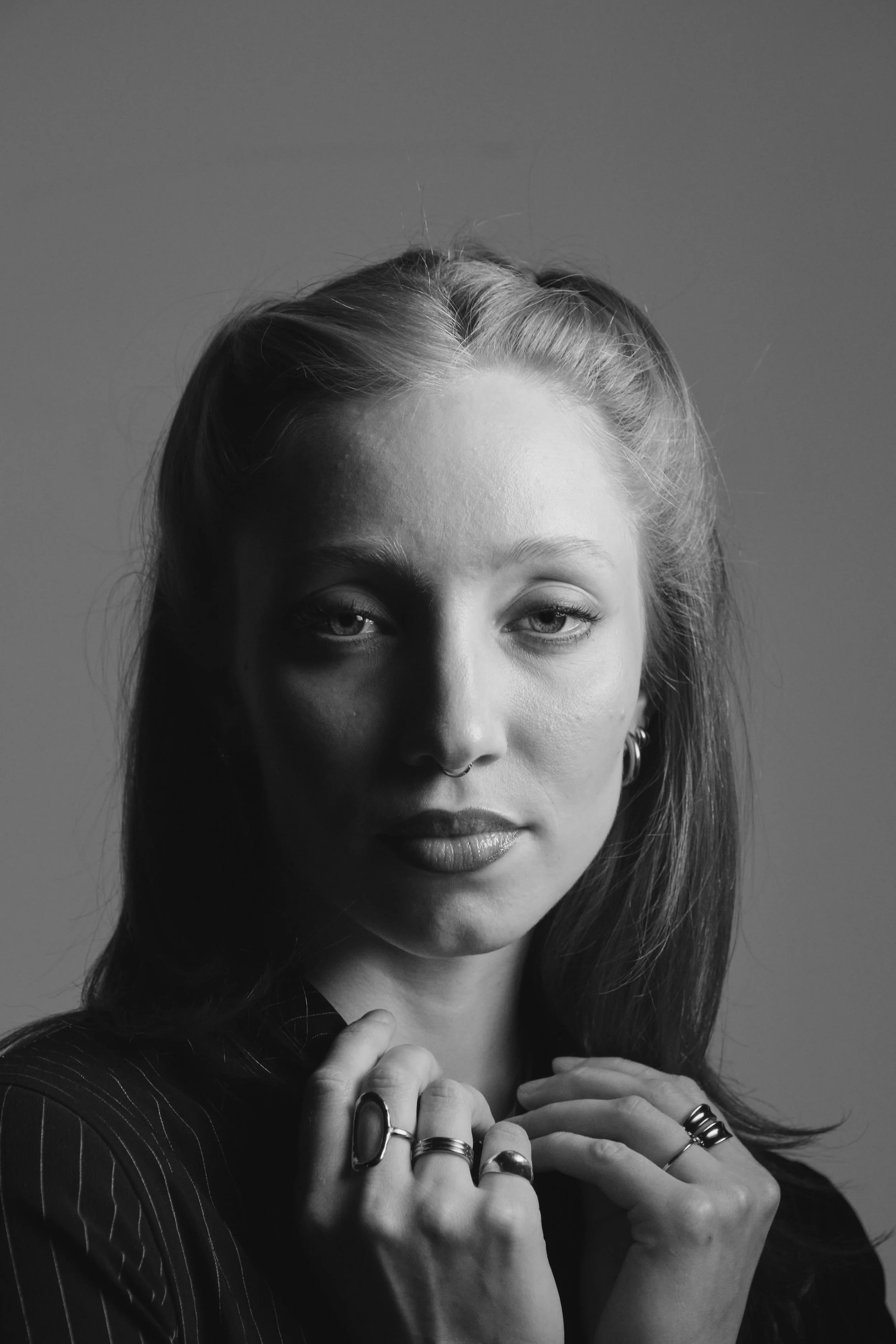 Schwarz-Weiß-Bild einer jungen Frau mit mehreren Ringen, make-up und einer Jacke mit Streifen, die direkt in die Kamera schaut.