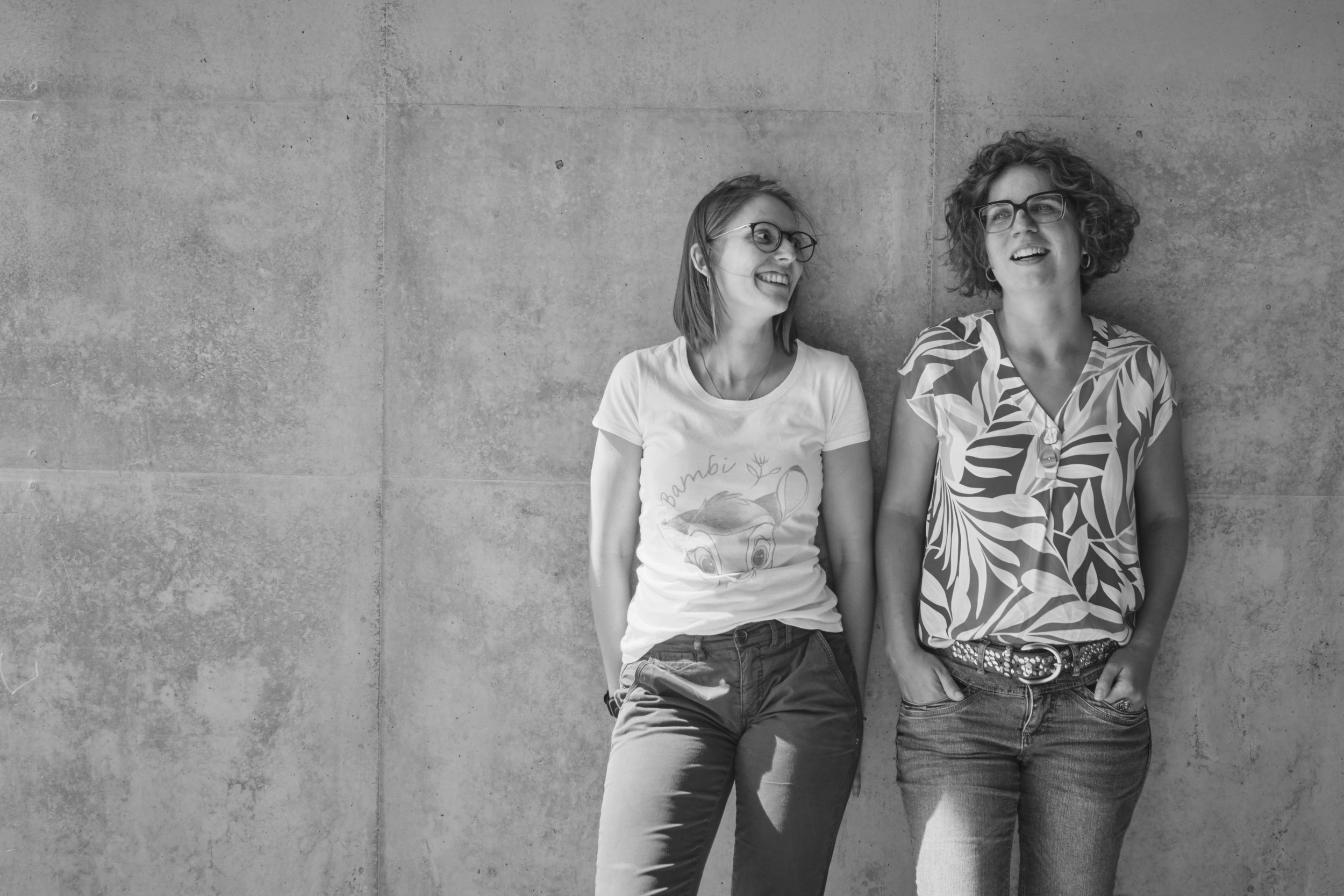 Zwei lachende Frauen stehen vor einer Betonwand, eine Frau trägt eine T-Shirt mit einem Eisbär und die andere trägt ein Hemd mit tropischem Muster.