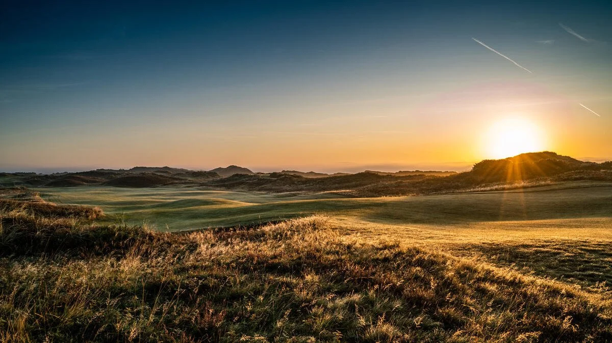 Sunrise over a coastal landscape with rolling hills and grassy terrain.