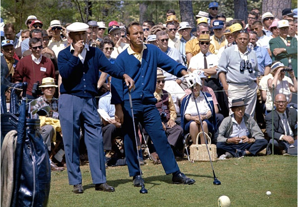Two men playing golf on a grass field while a large crowd watches.