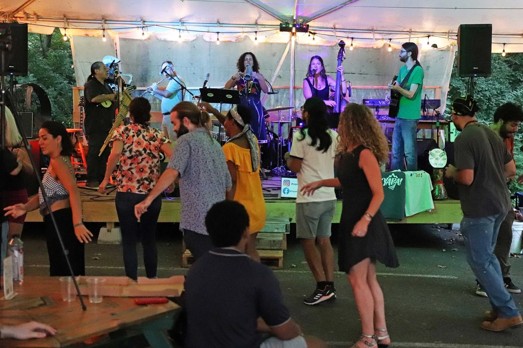 A live music band performs on stage at an outdoor event under string lights, with a dancing audience in front.