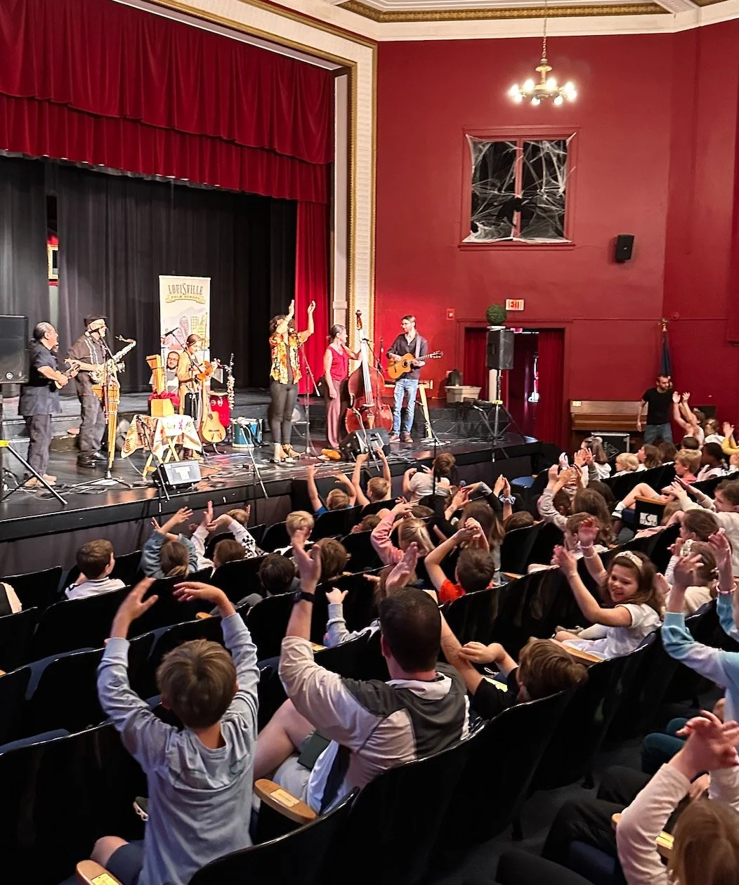 Children and adults watching a musical performance on stage with instruments, in a red and black theater with a red curtain and a decorative ceiling.