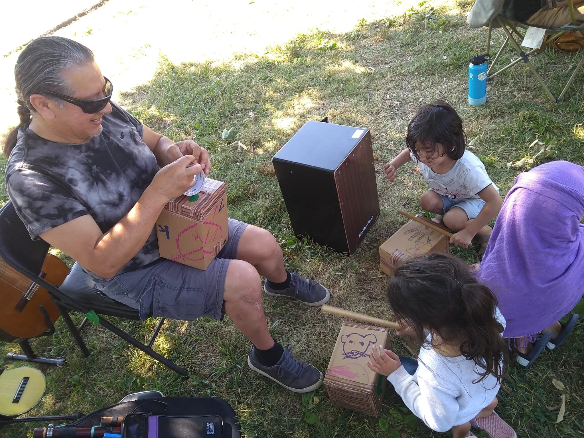 A man wearing sunglasses and a tie-dye shirt sitting outdoors on a black folding chair. He is smiling and holding a ring. Next to him are children drawing on cardboard boxes. There is green grass and trees providing shade, with a chair, water bottle, and guitar nearby.