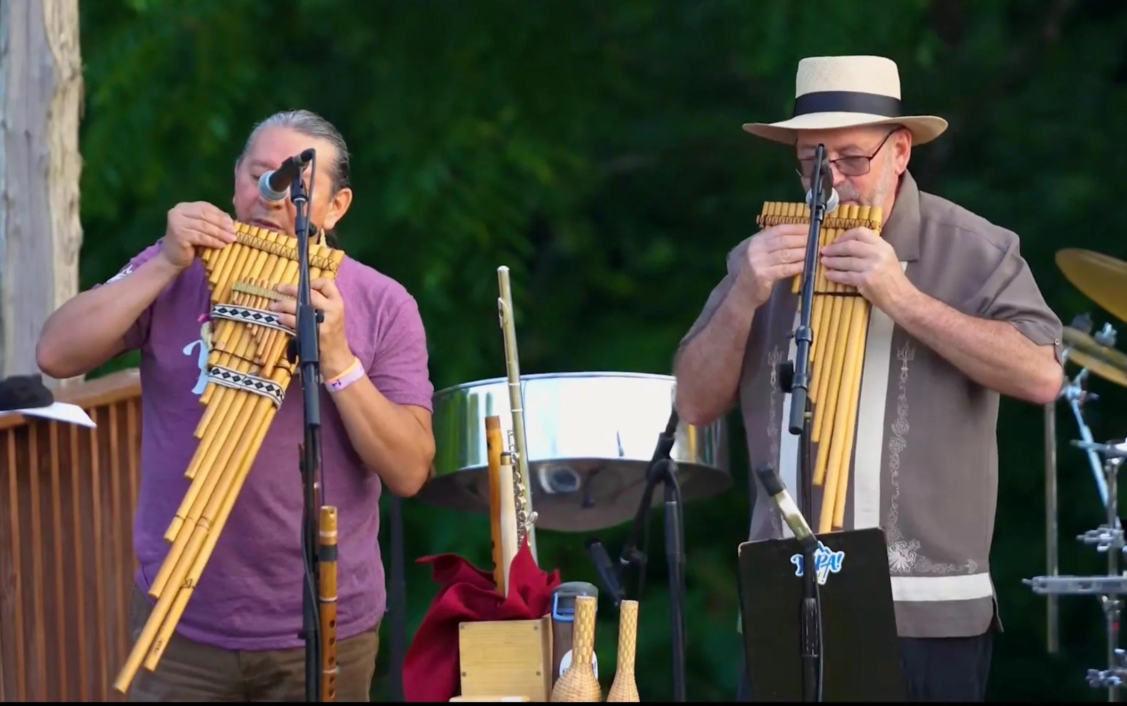 Two men playing pan flute instruments in an outdoor setting with trees in the background.