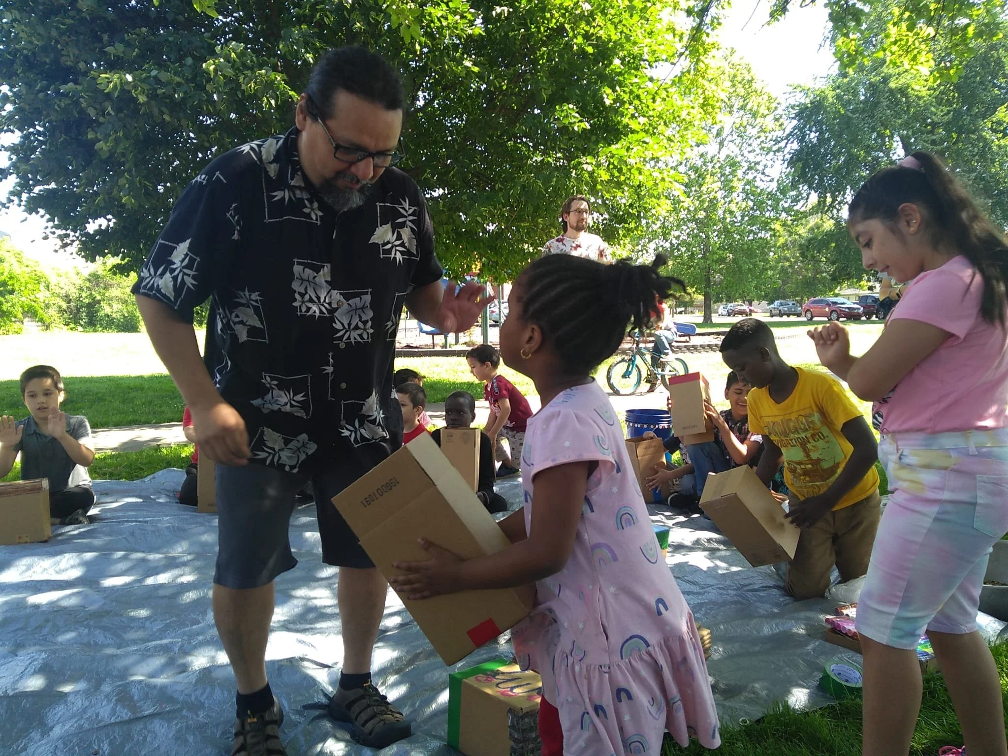 Children and adults gathered outdoors under a large tree during daytime, participating in an event where a man in a black Hawaiian shirt is handing out boxes to a girl in a pink dress, with other children waiting and adults in the background.