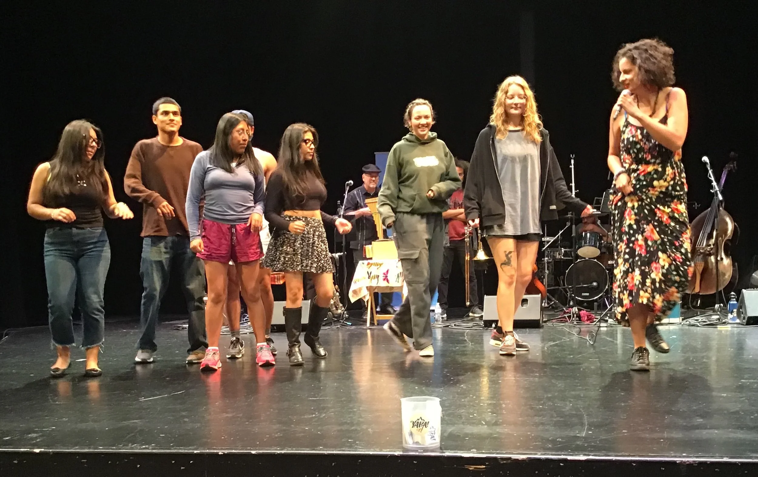 A group of diverse people standing on a stage, some wearing casual clothes, with a woman in a floral dress speaking into a microphone, and musical instruments in the background.