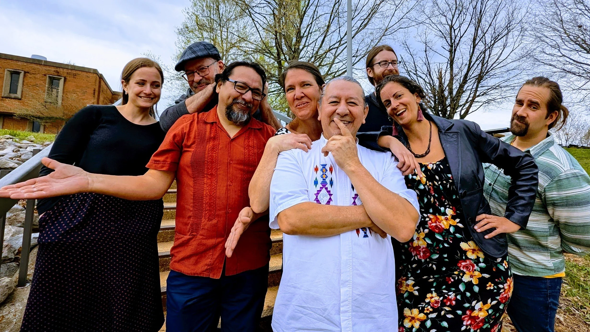 Group of eight diverse adults smiling and posing outdoors on steps with trees and a building in the background.