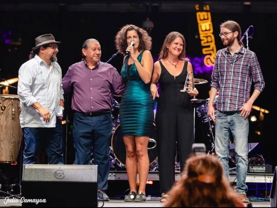 Group of five people standing on stage, with the woman in the middle holding a microphone and the woman next to her holding an award, at a music venue.