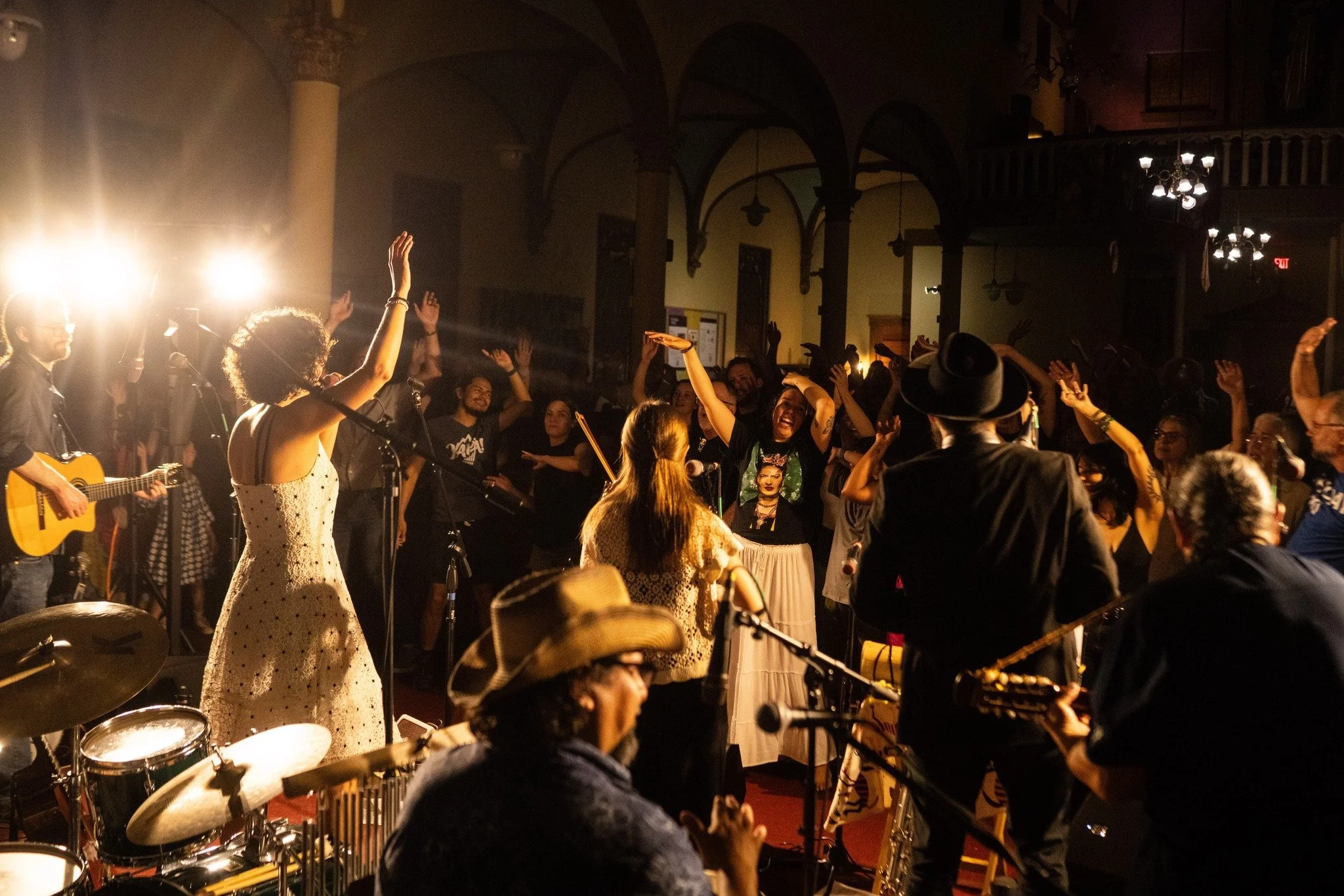 People dancing and singing at a lively indoor concert or event, with band members playing instruments including guitar and drums, warm lighting, and an ornate interior with arches and chandeliers.