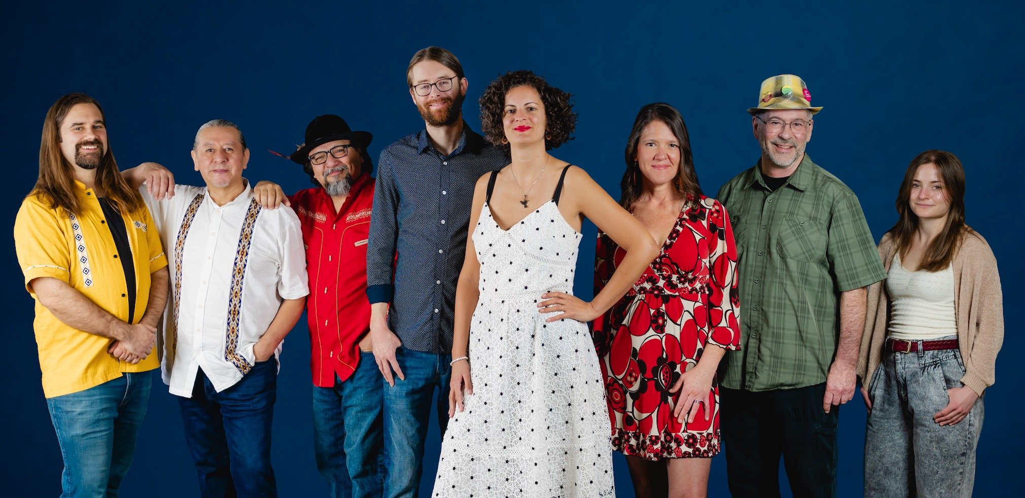 A diverse group of eight people standing together against a dark blue background, smiling and facing the camera.