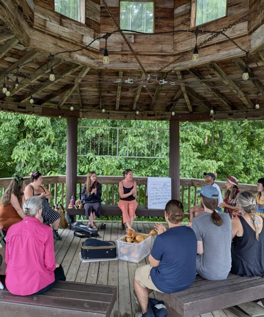 A group of people sitting on a wooden gazebo porch, attentively listening to a woman speaking. The gazebo is decorated with string lights and surrounded by green trees.