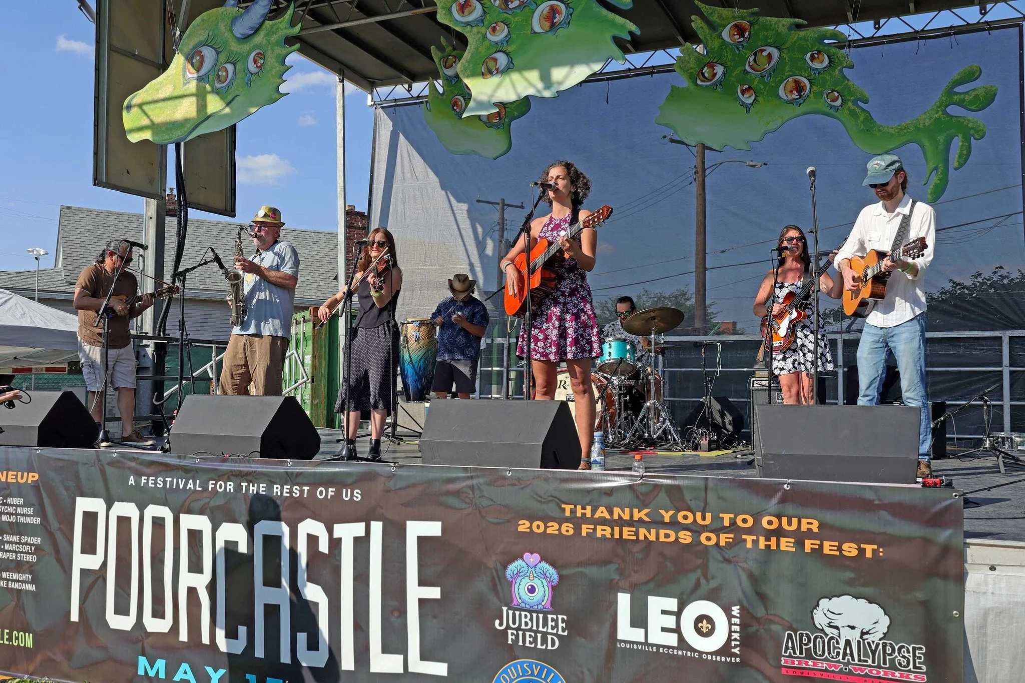 A group of musicians performing on an outdoor stage during a festival, with some playing guitars, a violin, drums, and a saxophone, under a colorful dragon-shaped banner.