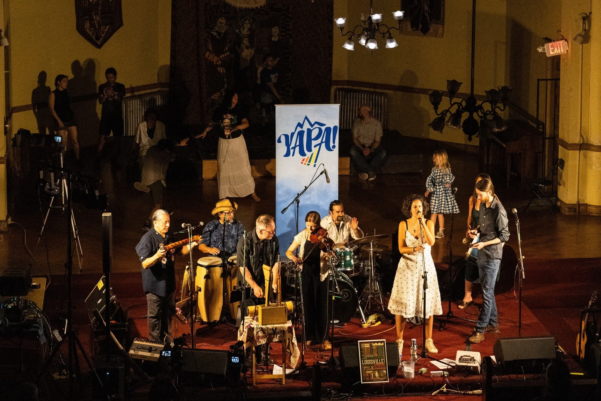 A live music performance at Louisville Folk School featuring musicians playing various instruments while a woman sings into a microphone. Several people are sitting and standing around on stage. A large sign reading 'YAPA!' and a poster for Louisville Folk School are visible, with a yellow wall and chandelier in the background.