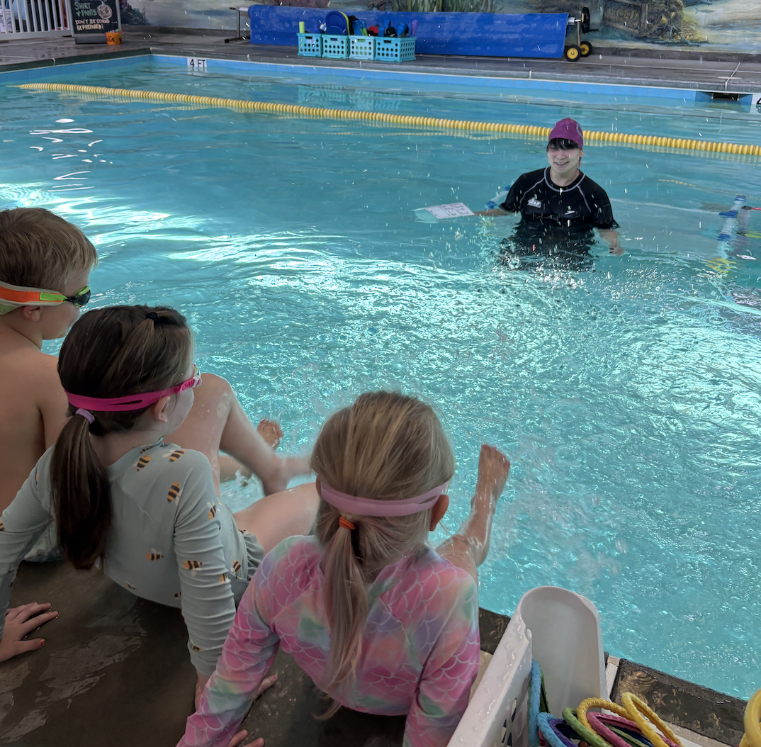 Children sitting by the edge of an indoor swimming pool, watching a swimming instructor, who is standing in the water, giving a lesson.