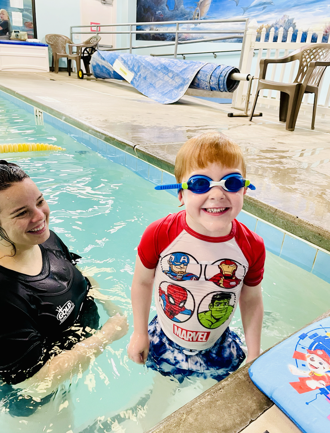 A young boy with red hair, wearing Marvel superhero swim shirt and blue goggles, smiling in a swimming pool with an adult woman nearby.