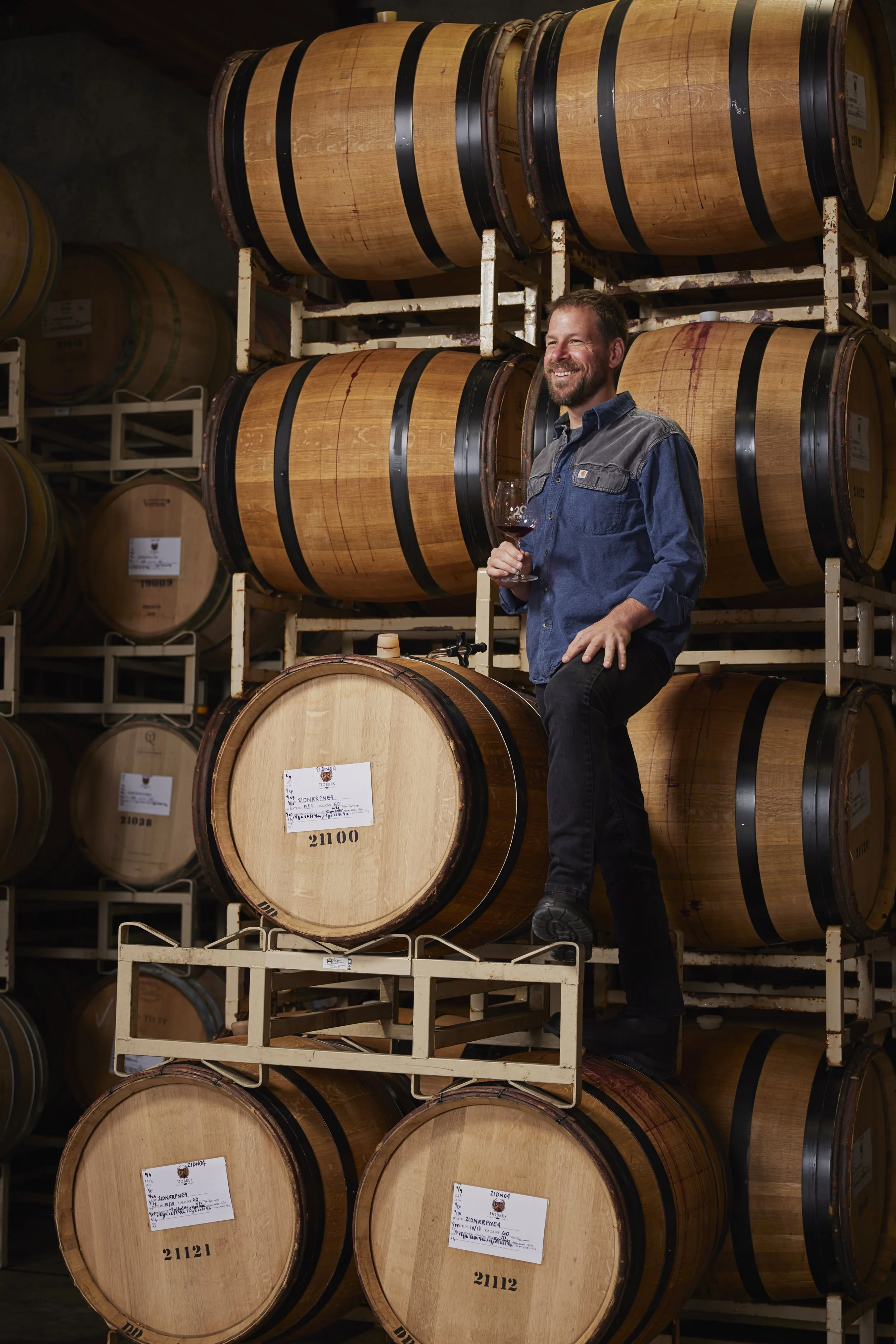 A man holding a glass of red wine, standing on a raised platform among large wooden wine barrels in a cellar.