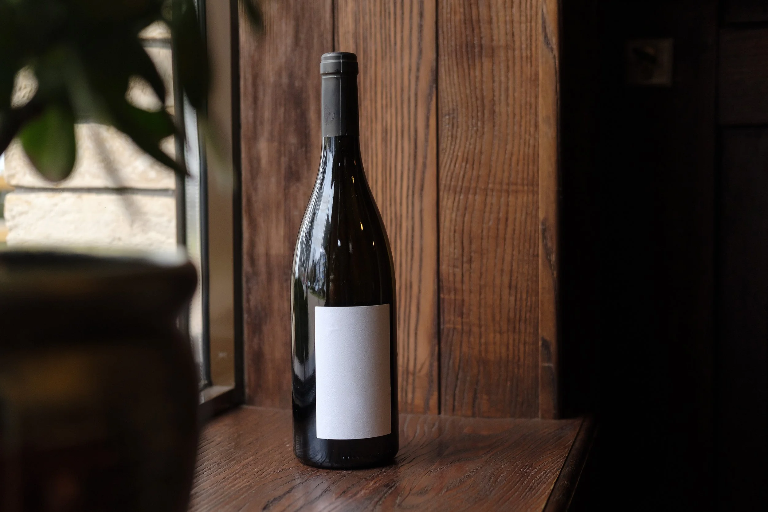 A dark wine bottle with a blank white label on a wooden surface near a window, with a blurred plant and pot in the foreground.
