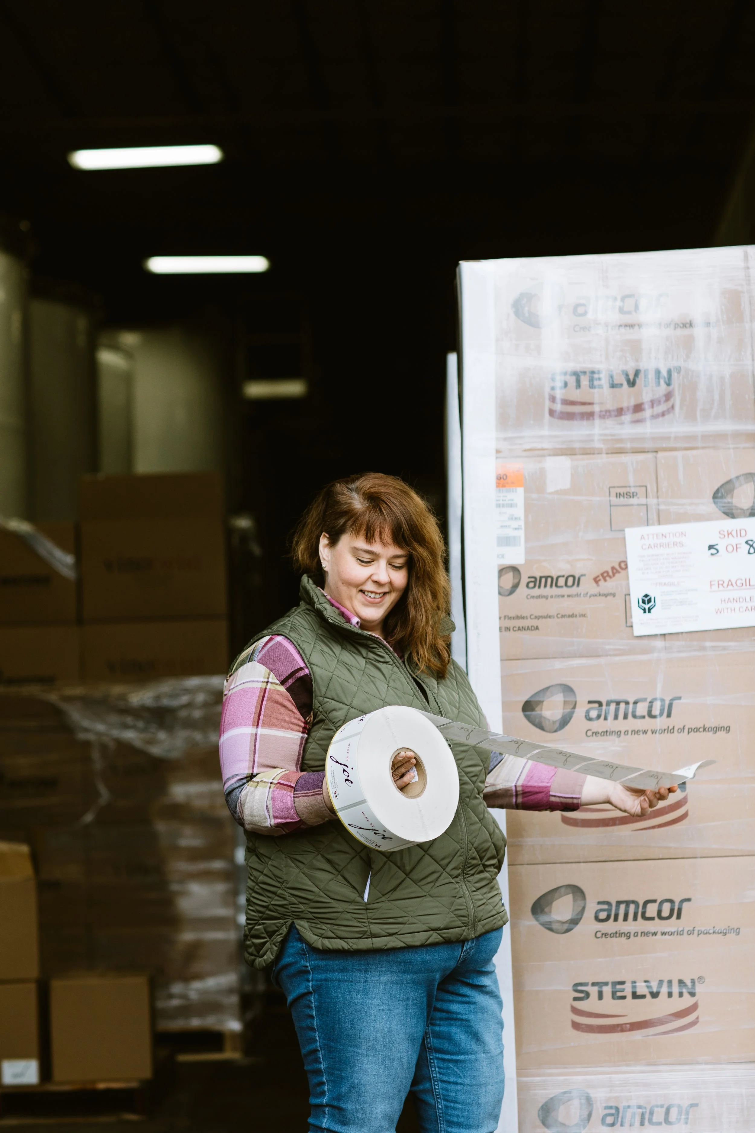 A woman with red hair wearing a green quilted vest and a plaid shirt is smiling while holding a roll of packaging tape and a strip of tape in a warehouse surrounded by large boxes.