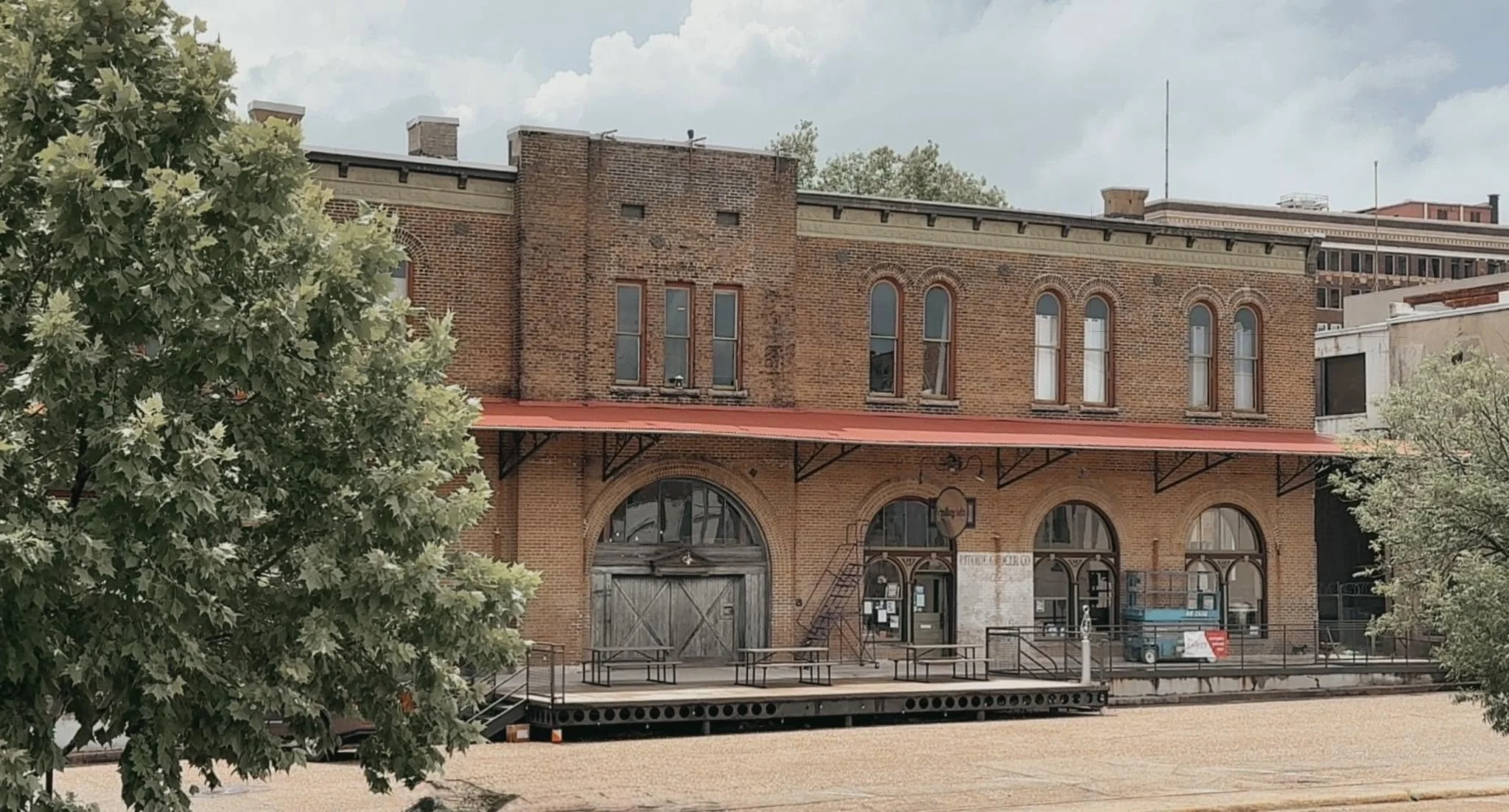 Historic brick building with large arched windows, a red awning, and a small tree in the foreground.