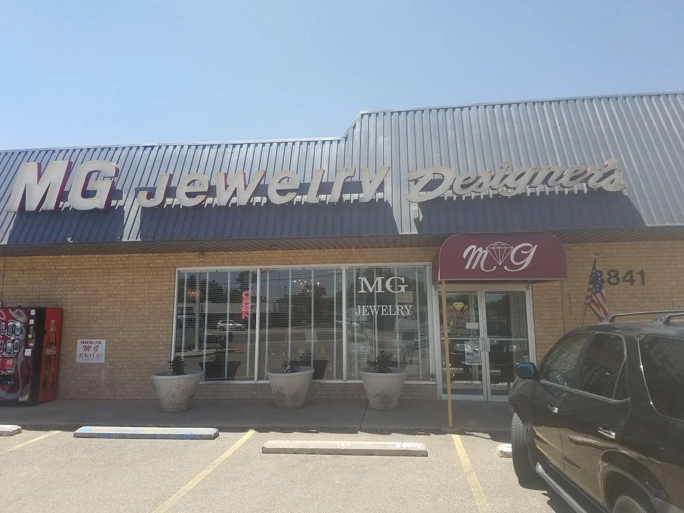 Exterior of MG Jewelry store with large sign reading 'M.G Jewelry Designers' on a metal awning, brick building entrance with glass windows, potted plants, and parking spots in front.