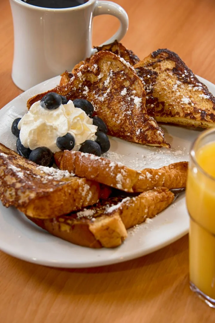 A breakfast plate with French toast topped with powdered sugar, a dollop of whipped cream with blueberries, a cinnamon stick, and a cup of coffee. A glass of orange juice is partially visible in the foreground.