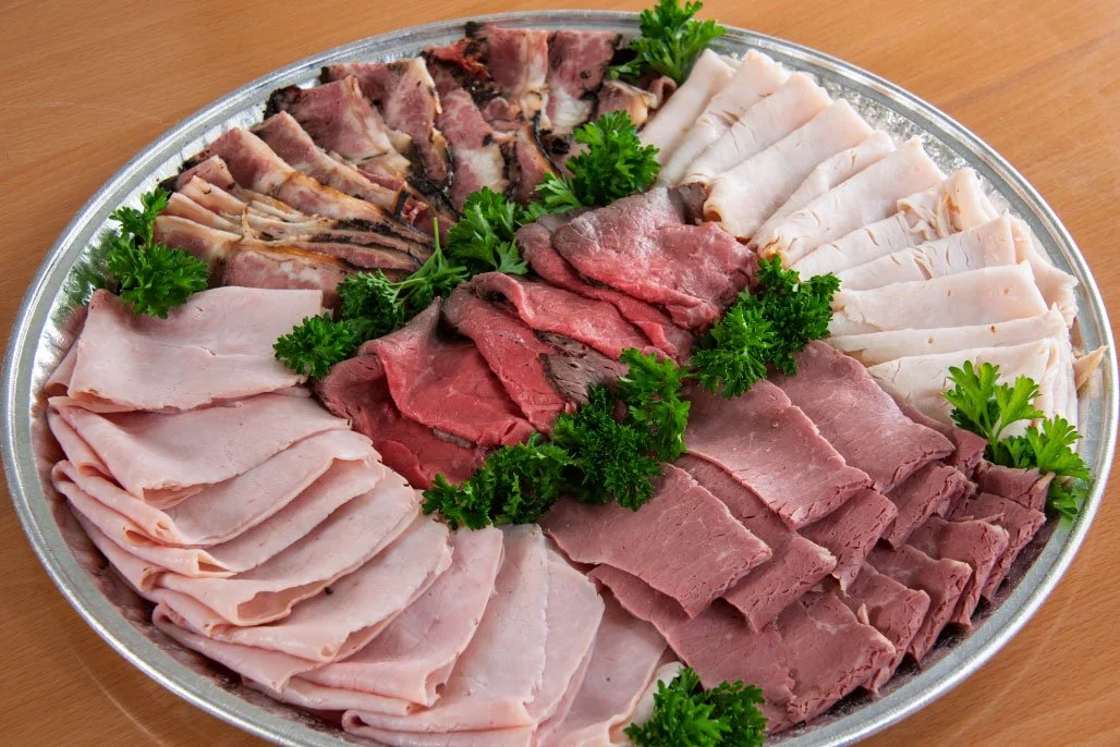 A large round tray of assorted cold cuts garnished with parsley, including slices of turkey, ham, roast beef, and other deli meats.
