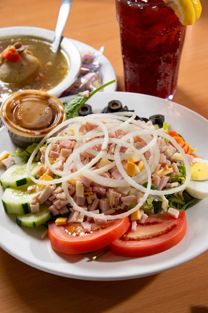 A seafood salad with sliced tomatoes, cucumbers, onions, shredded cheese, and chopped ham on a white plate, accompanied by a small container of dressing. In the background, there is a bowl of soup and a glass of dark soda with lemon wedges.