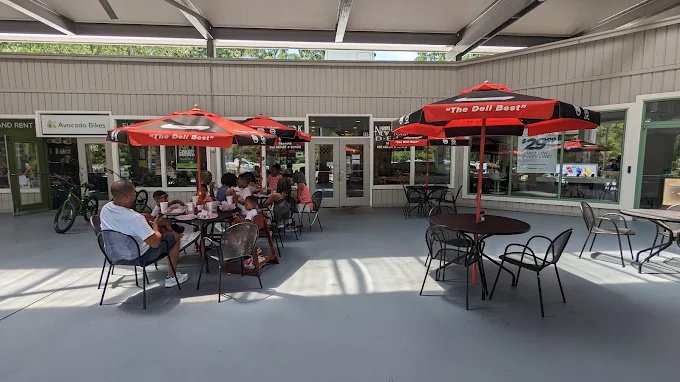 Outdoor patio area with red umbrellas, tables, and chairs at a restaurant or cafe, with people sitting and dining.