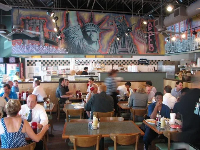 Inside a busy restaurant with many patrons seated at wooden tables, a large mural of the Statue of Liberty and other New York landmarks decorates the back wall, with an 'ARIBO' sign hanging vertically.