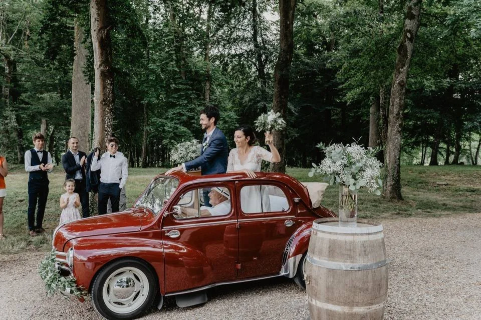 Un couple en tenue de mariage avec des invités en arrière-plan, dans une forêt, avec une voiture ancienne rouge décorée pour un mariage.