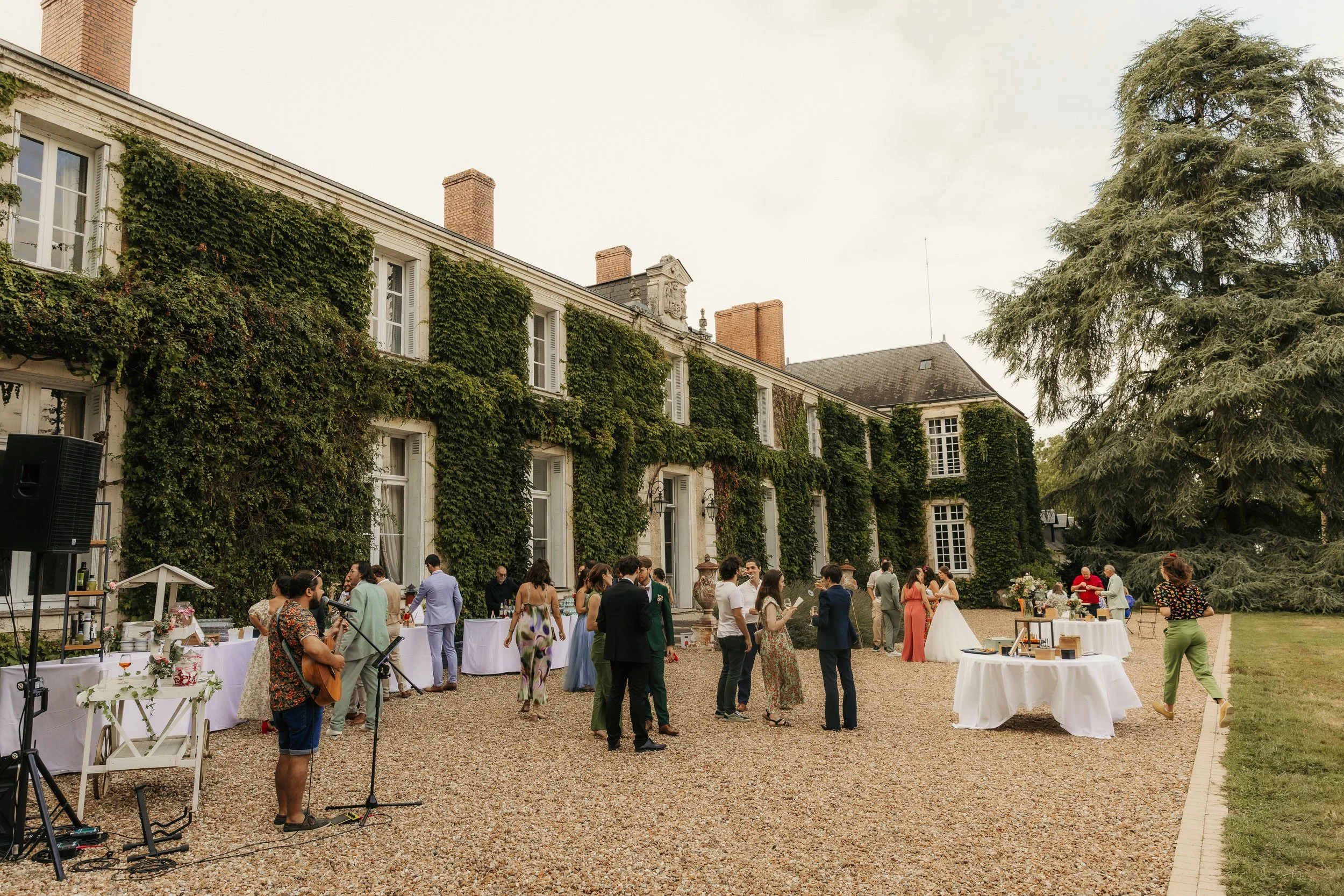 Célébration en extérieur devant un château avec des invités, un musicien jouant de la guitare, tables de nourriture, et un groupe de personnes discutant à côté d'une grande maison couverte de lierre.