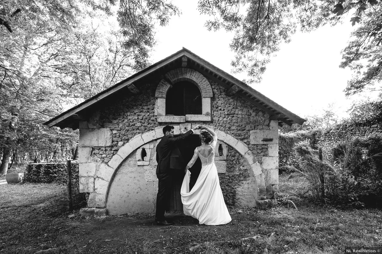 Un couple en robe de mariée et costume dansant devant une petite vieille maison en pierre, entourée d'arbres, dans une forêt.