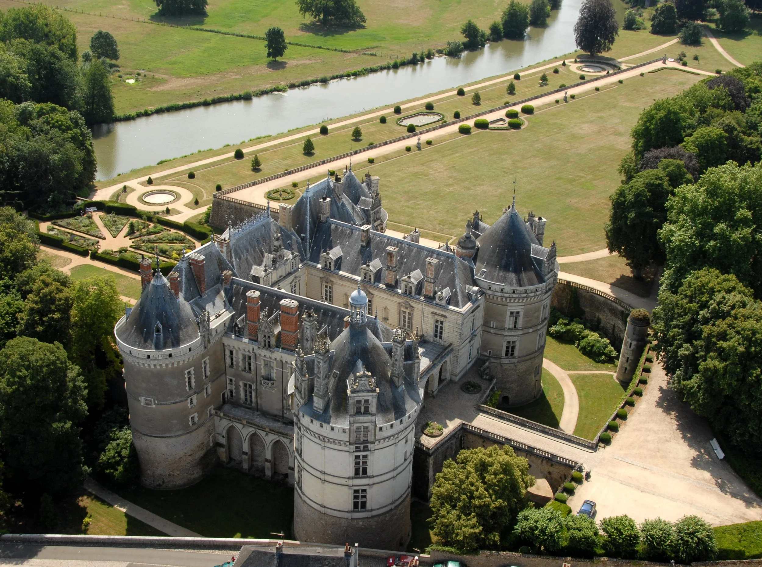 Château médiéval entouré de jardins verdoyants, avec une allée menant à l'entrée principale, et une rivière en arrière-plan.